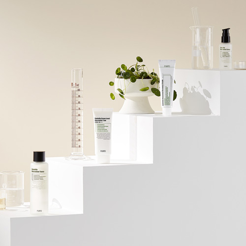 푸리토 센텔라 제품들이 계단 형태로 배치되어 있는 연출 사진 Purito's Centella skincare line arranged on white steps with lab glassware and green plants