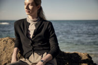 바닷가 바위 위에 앉아 카디건과 라이트한 이너를 입은 여성 모델 Woman seated on a coastal rock wearing a black cardigan over a light inner top