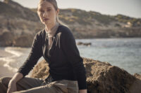 바닷가 바위 위에 앉아 블랙 카디건과 시어한 이너를 레이어드한 여성 모델 Woman seated on a rock by the sea wearing a dark cardigan over a sheer inner top