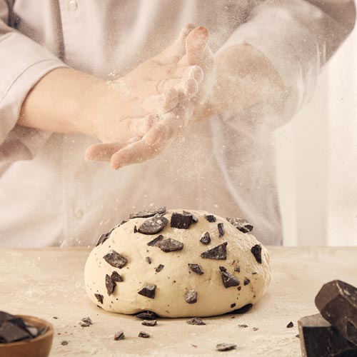 Baker clapping flour above chocolate chip dough on wooden table