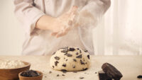 Baker clapping flour above chocolate chip dough on wooden table