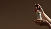 두 손으로 젠틀 블랙 클렌징 오일을 들어올린 장면 Hands holding a Gentle Black Cleansing Oil against a brown background