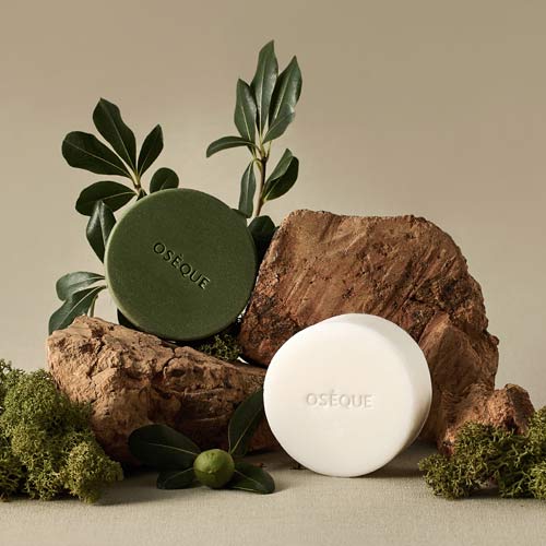 초록과 흰색의 오제끄 천연비누가 자연 요소들과 함께 배치된 제품컷 Two natural soaps from Oseque arranged with moss, leaves, and stone elements in a warm-toned setting