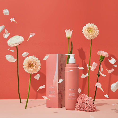 꽃잎이 흩날리는 배경에 배치된 오제끄 복숭아꽃 바디로션 Peach-colored body lotion bottle with scattered flower petals in the background