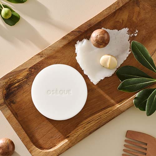 나무 트레이 위에 놓인 오제끄 화이트 천연비누와 맥아다미아 오일, 나뭇잎, 나무빗 A white Oseque natural soap on a wooden tray with macadamia nuts, oil texture, and leaves