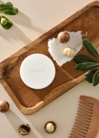 나무 트레이 위에 놓인 오제끄 화이트 천연비누와 맥아다미아 오일, 나뭇잎, 나무빗 A white Oseque natural soap on a wooden tray with macadamia nuts, oil texture, and leaves