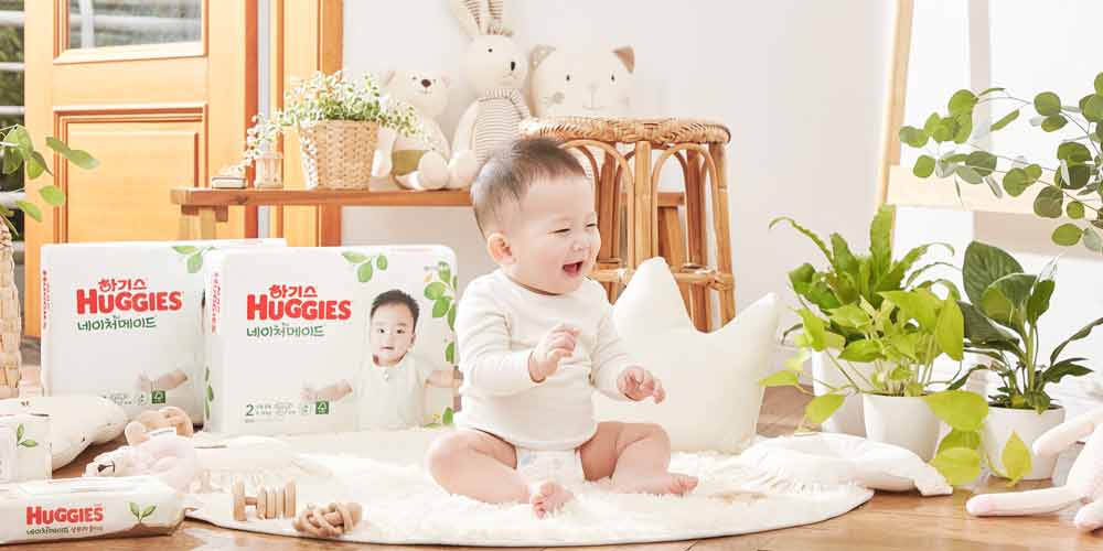 밝은 자연광이 드는 실내에서 웃고 있는 아기와 하기스 네이처메이드 기저귀 제품들 Huggies Nature Made diaper packaging with a smiling baby in a plant-filled, naturally lit interior