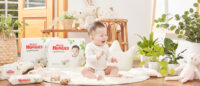 밝은 자연광이 드는 실내에서 웃고 있는 아기와 하기스 네이처메이드 기저귀 제품들 Huggies Nature Made diaper packaging with a smiling baby in a plant-filled, naturally lit interior