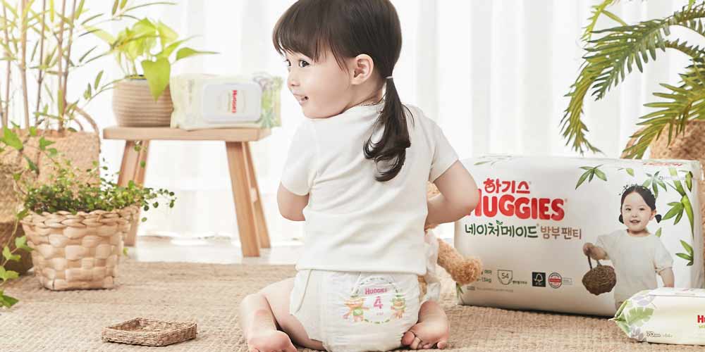 기저귀를 착용한 여자아이가 뒤를 돌아보며 웃고 있고, 옆에는 하기스 밤부 기저귀 패키지와 물티슈가 함께 배치되어 있음 Toddler girl turning back and smiling while wearing Huggies diaper, next to a product pack and baby wipes