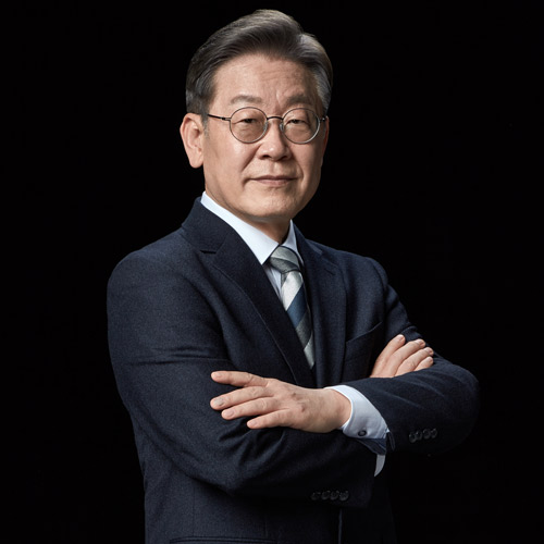 검은 배경 앞에서 팔짱을 끼고 카메라를 응시하는 이재명 대통령 후보의 전신 포트레이트 A full-length portrait of presidential candidate Lee Jae-myung standing with arms crossed, facing the camera against a black background