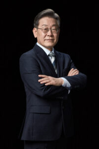 검은 배경 앞에서 팔짱을 끼고 카메라를 응시하는 이재명 대통령 후보의 전신 포트레이트 A full-length portrait of presidential candidate Lee Jae-myung standing with arms crossed, facing the camera against a black background. Korean presidential election photo