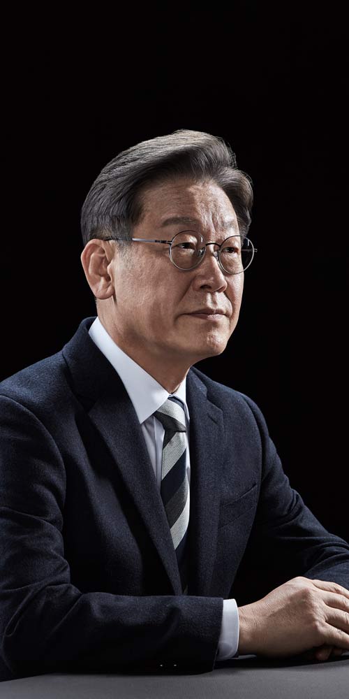 정면에 가까운 앵글로 테이블 앞에 앉아 단정한 자세를 취한 이재명 대통령 후보의 포트레이트 A composed and front-facing portrait of presidential candidate Lee Jae-myung seated at a table with a calm demeanor