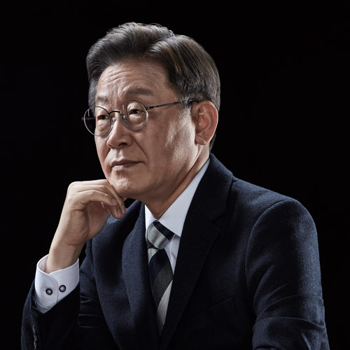 검은 배경 앞 테이블에 팔을 올리고 앉아 진중한 표정을 짓고 있는 이재명 대통령 후보의 공식 포트레이트 A formal portrait of presidential candidate Lee Jae-myung seated at a table with a contemplative expression against a black background