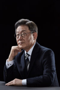 검은 배경 앞 테이블에 팔을 올리고 앉아 진중한 표정을 짓고 있는 이재명 대통령 후보의 공식 포트레이트 A formal portrait of presidential candidate Lee Jae-myung seated at a table with a contemplative expression against a black background. Korean presidential election photo