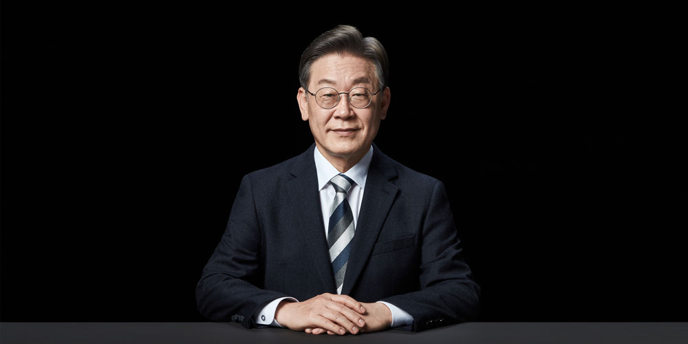 검은 배경 앞 테이블에 앉아 정면을 바라보며 미소 짓는 이재명 대통령 후보의 포트레이트 A front-facing portrait of presidential candidate Lee Jae-myung seated at a table, smiling slightly against a black background