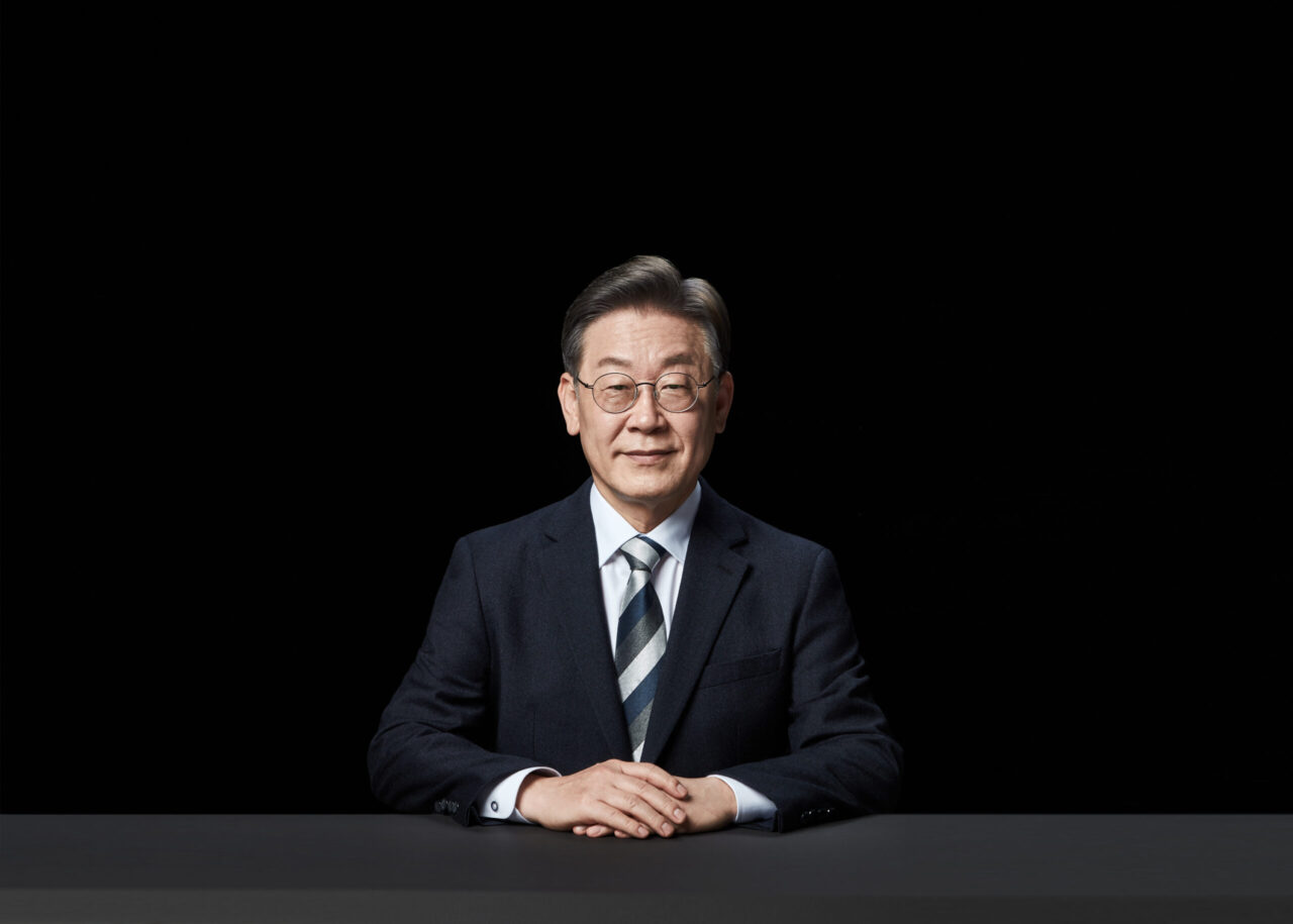 검은 배경 앞 테이블에 앉아 정면을 바라보며 미소 짓는 이재명 대통령 후보의 포트레이트 A front-facing portrait of presidential candidate Lee Jae-myung seated at a table, smiling slightly against a black background, Korean presidential election photo