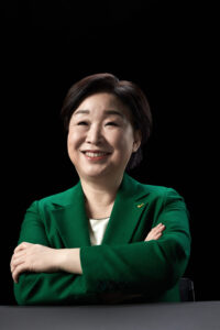 검은 배경 앞에서 팔짱을 낀 채 미소 짓는 심상정 대통령 후보의 공식 포트레이트 A confident smiling portrait of presidential candidate Shim Sang-jung with arms crossed against a black background