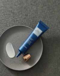 회색 원형 접시 위에 놓인 오제끄 블루 튜브형 크림과 질감 샘플 A blue tube of Oseque facial cream with a smear of product on a grey dish