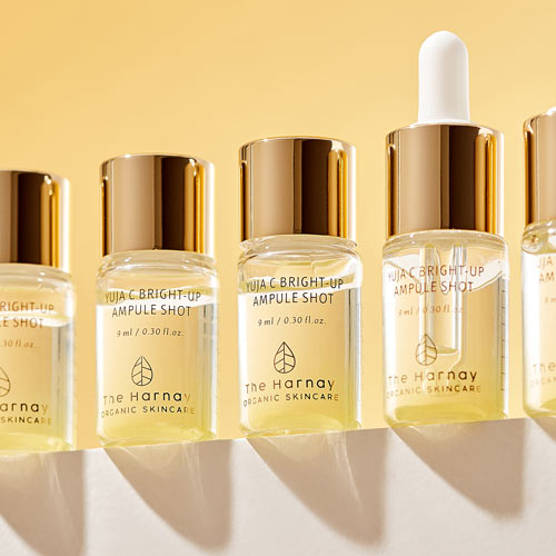 황금빛 배경 앞에 줄지어 선 유자C 브라이트 업 앰플 제품 이미지 Lineup of The Harnay’s Yuja C ampoule bottles under golden light