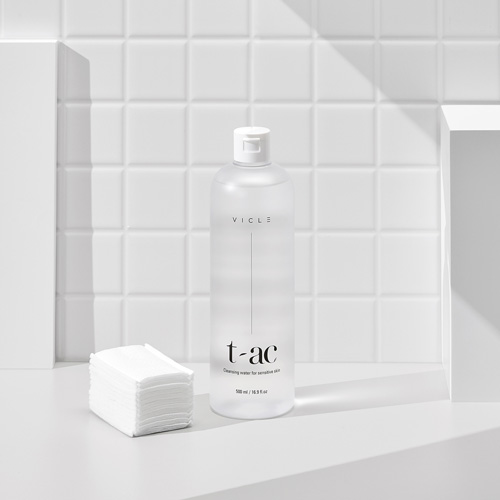 화이트 타일 벽 앞에 클렌징 워터와 화장솜이 나란히 놓인 구성 A clear Vicle cleansing water bottle and stacked cotton pads set against white bathroom tiles
