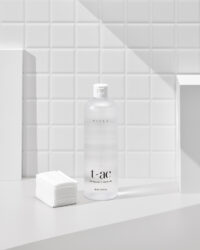 화이트 타일 벽 앞에 클렌징 워터와 화장솜이 나란히 놓인 구성 A clear Vicle cleansing water bottle and stacked cotton pads set against white bathroom tiles