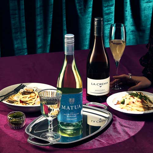 두 병의 화이트 와인과 파스타 요리가 어우러진 정갈한 테이블 연출 이미지 A composed table setting with two bottles of white wine, pasta dishes, and elegant props on deep-toned fabric