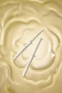 골드 제형 위에 놓인 제나셀 주사기형 세럼 제품 사진 JenaCell syringe-type serum placed on golden textured liquid background